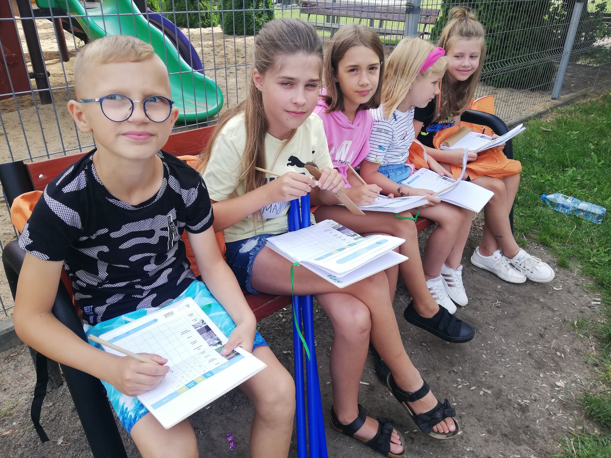 Plener, uczestnicy Warsztatów z Klimatem na ławeczce przy placu zabaw, wypełniają karty zadań Plac zabaw za biblioteką, ławka, siatka, piasek, zjeżdżalnia, karty pracy, długopisy, dzieci, trawa